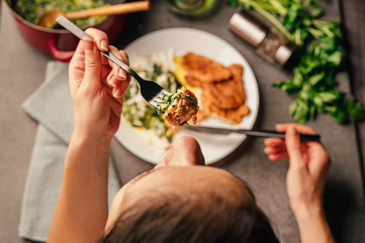 Person eating chicken with a fork, plate of food in front including a piece of chicken and side vegetables